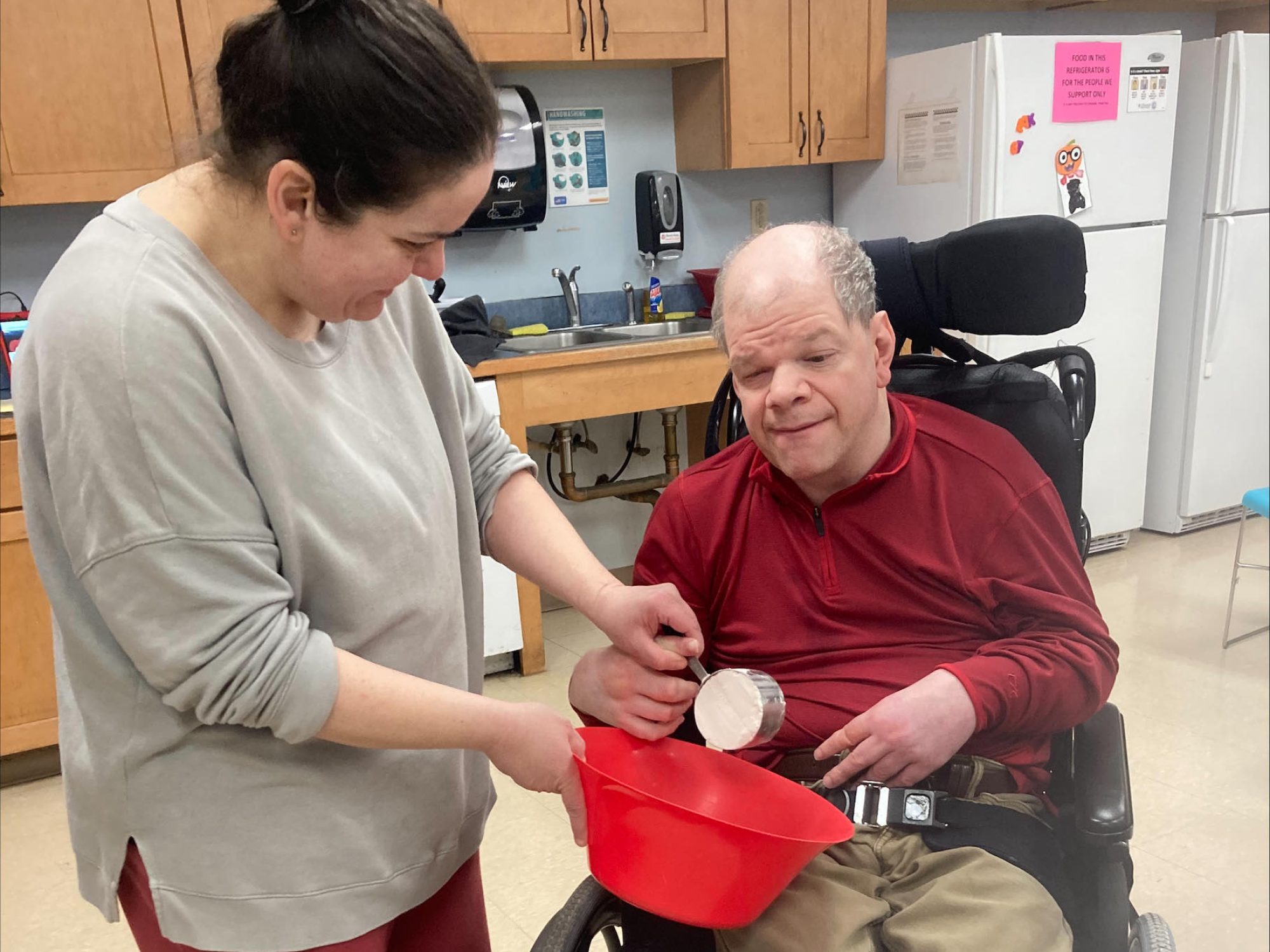 A staff member assists a program participant in a wheelchair with a cooking activity in a community day program kitchen