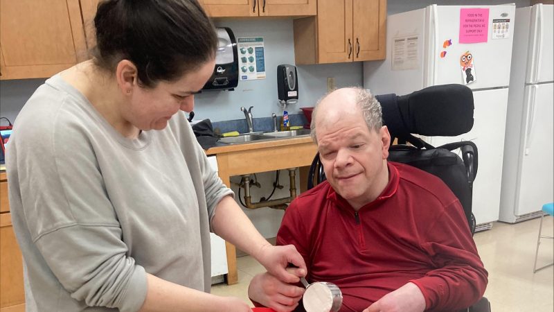 A staff member assists a program participant in a wheelchair with a cooking activity in a community day program kitchen