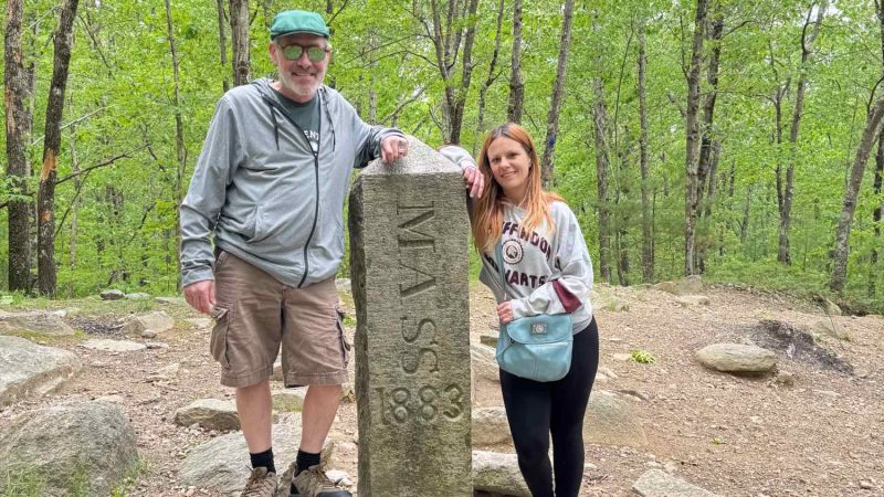 Two people posing next to a Massachusetts state boundary marker during an outdoor community outing
